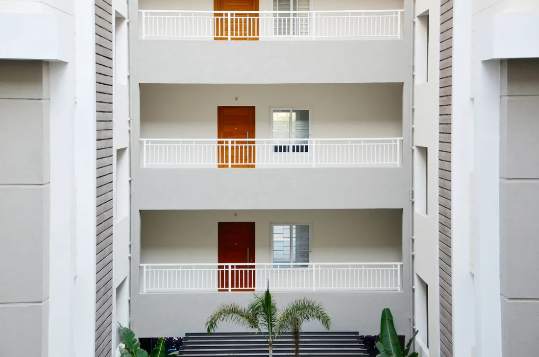Façade blanche d'un immeuble de logements sur trois niveaux, vue depuis le sol : coursives à garde-corps blancs, portes en bois et palmiers en pied de bâtiment
