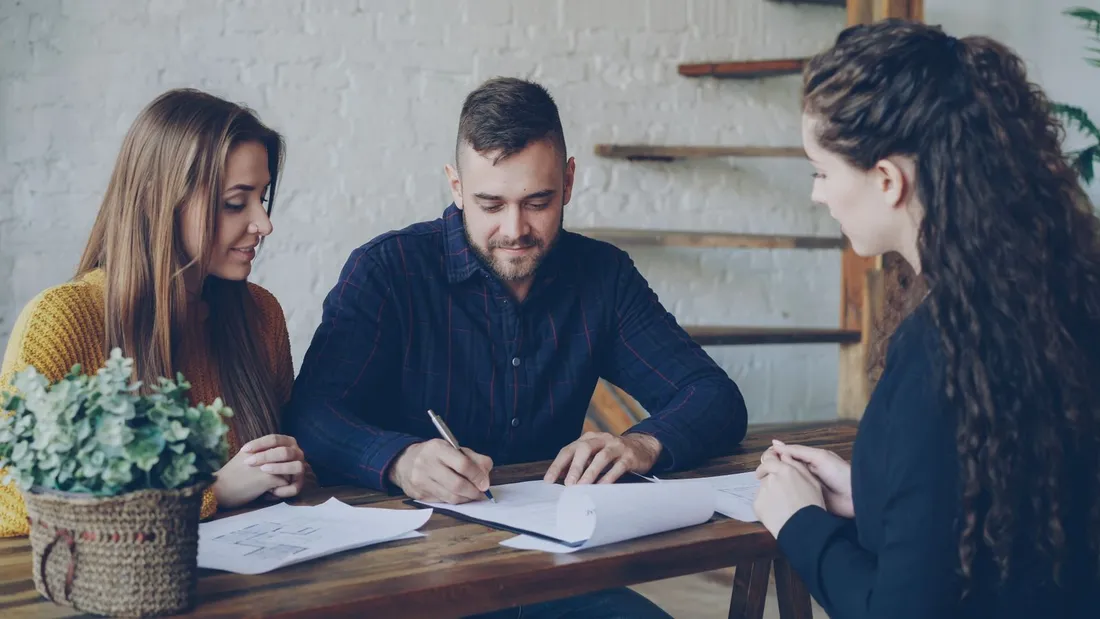 Un homme signe un document posé sur une table en bois, une femme à sa gauche et une conseillère face à eux, plans de logement étalés sur la table