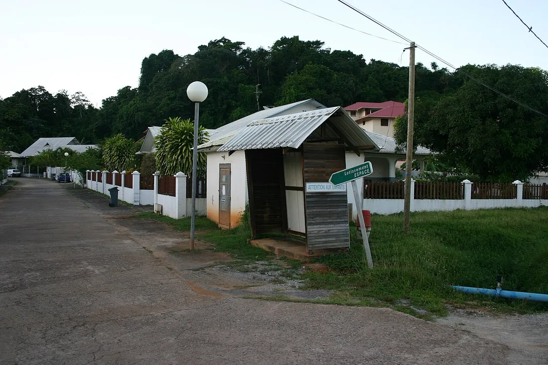 Rue d'un lotissement à Cayenne, en Guyane : chaussée en béton, abribus en bois et tôle, panneau « Lotissement Espace », maisons basses derrière des clôtures et végétation dense à l'arrière-plan