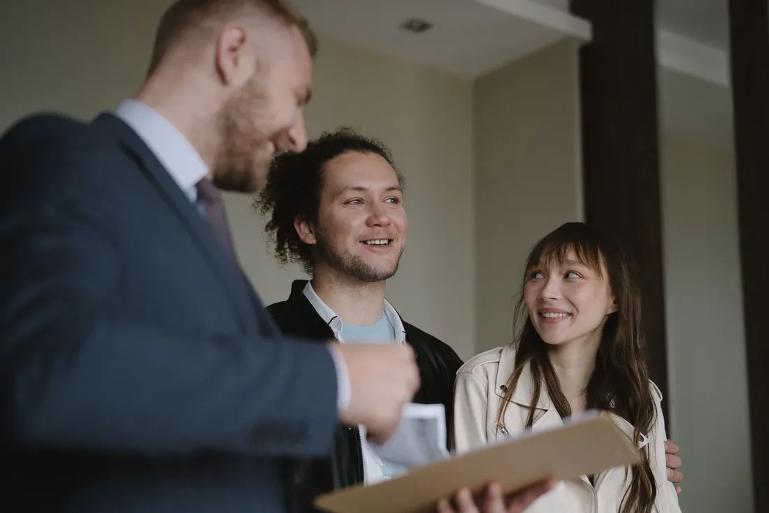 Un agent immobilier en costume, un dossier à la main, discute avec un jeune couple dans le hall d'un immeuble d'habitation