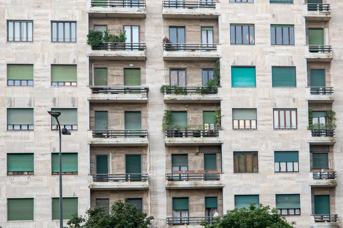 Façade d'un immeuble d'habitation collectif vue depuis la rue : balcons superposés, volets baissés et plantes en pot devant les logements