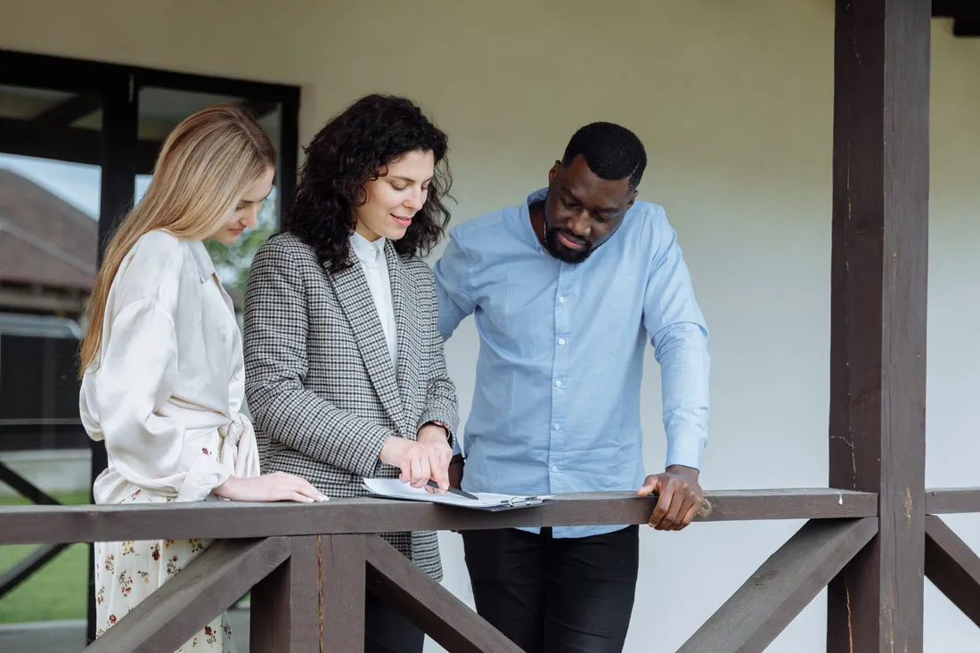 Une conseillère en gestion locative commente un document posé sur la rambarde d'une terrasse, entourée d'un couple de propriétaires attentifs