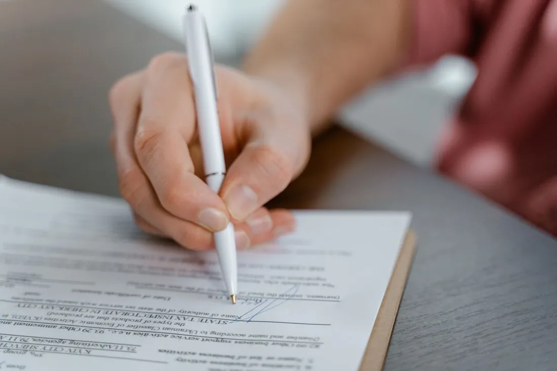 Gros plan sur une main qui signe au stylo le bas d'un document administratif posé sur un bureau
