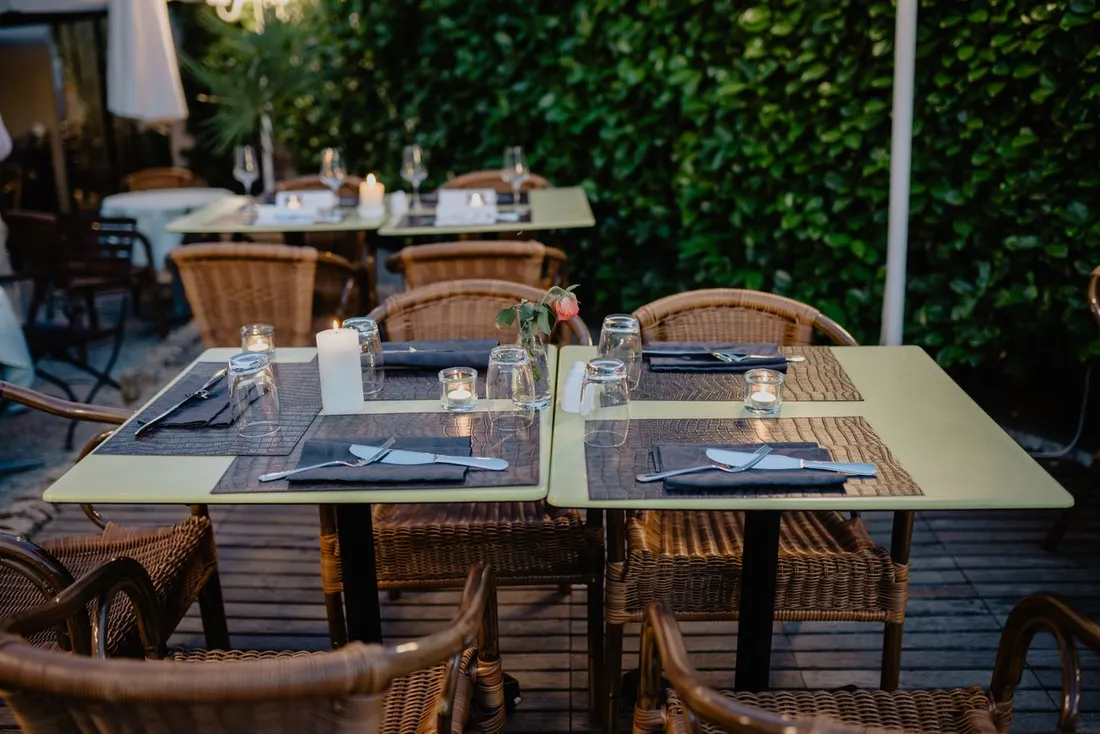 Terrasse de restaurant dressée pour le dîner, tables avec bougies, chemins de table et fauteuils en rotin devant un mur de verdure