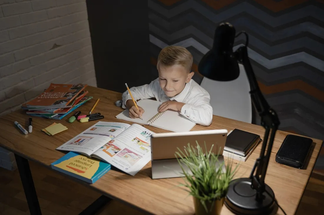 Jeune enfant faisant ses devoirs à un bureau avec cahiers, manuels et tablette sous une lampe