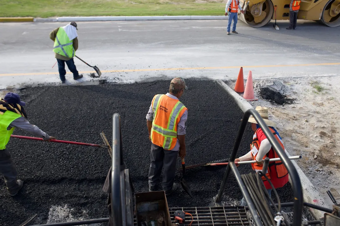 Ouvriers en gilets de sécurité étalant à la pelle et au râteau de l'enrobé noir sur une chaussée, un compacteur et des cônes de chantier à l'arrière-plan