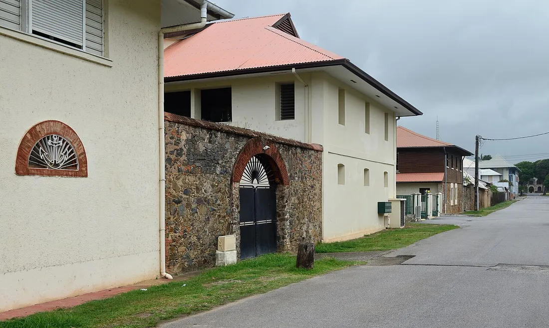 Rue de Cayenne, en Guyane, vue depuis le sol : bâtiments clairs à toiture rouge, mur de moellons percé d'un portail cintré, motif d'ancre dans un tympan, chaussée étroite bordée d'herbe