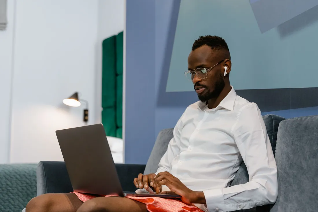 Professionnel en chemise blanche installé sur un canapé et travaillant sur son ordinateur portable dans un logement meublé, typique d'un séjour d'affaires de plusieurs semaines