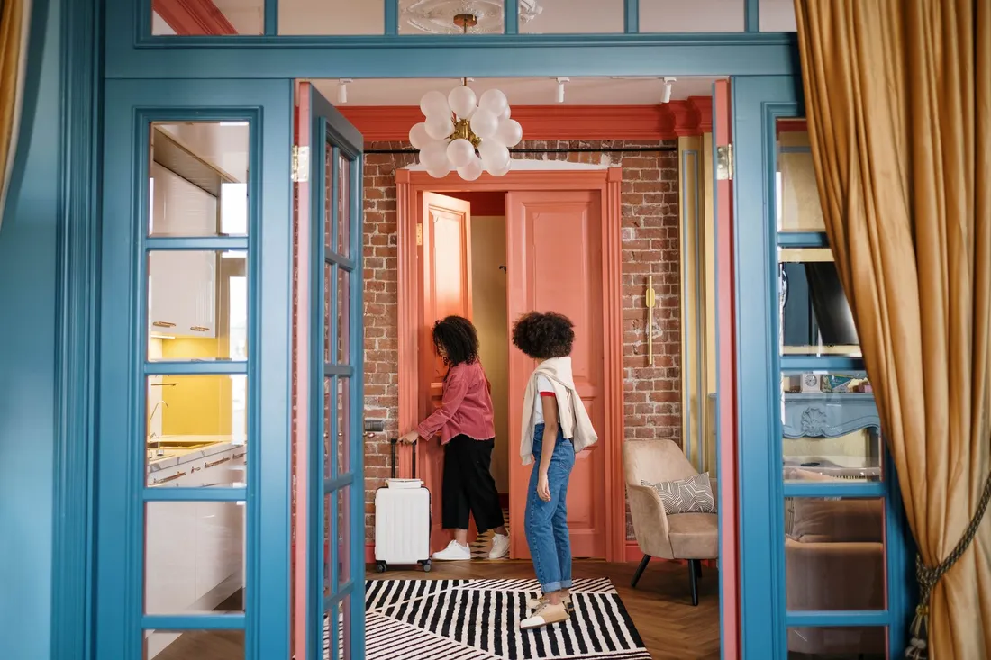 Deux femmes franchissent la porte d'un appartement meublé, l'une tirant une valise à roulettes derrière elle