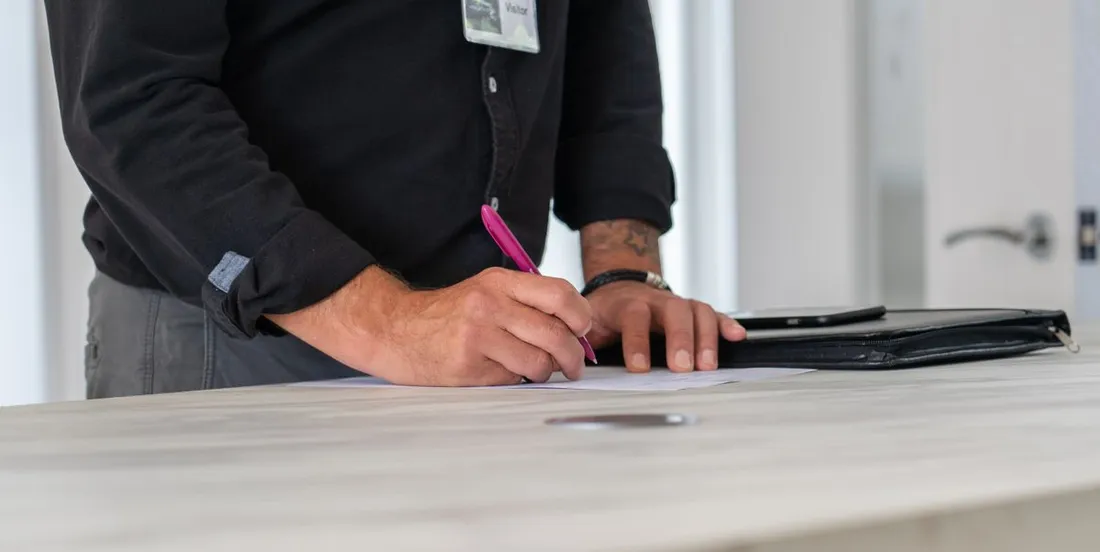 Gros plan sur les mains d'un homme portant un badge visiteur, qui remplit un formulaire au stylo sur un comptoir clair, une pochette à documents posée à côté
