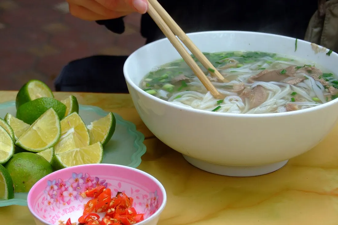 Grand bol de soupe chinoise fumante aux nouilles de riz et bouillon clair, servi avec baguettes, quartiers de citron vert et piments frais