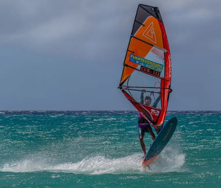 Windsurfeur en plein planning sur une mer turquoise, voile colorée déployée et gerbe d'écume, scène évoquant les spots de windsurf de l'Atlantique