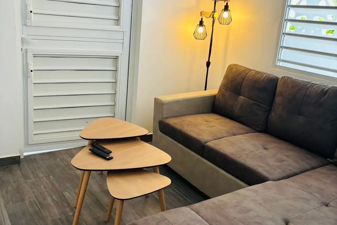 Séjour de la maison Tropikana : canapé d’angle brun, tables gigognes en bois clair et lampadaire à ampoules apparentes