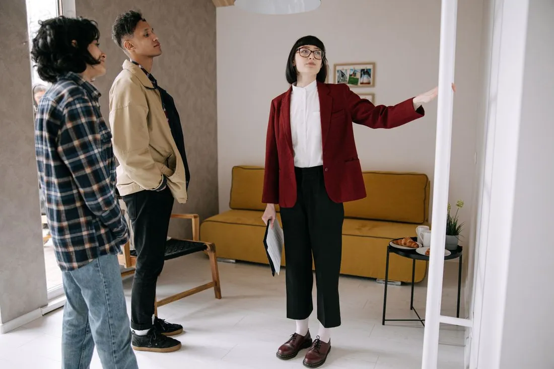 Une agente immobilière, dossier à la main, présente une pièce d'appartement à un couple de visiteurs debout dans l'encadrement de la porte