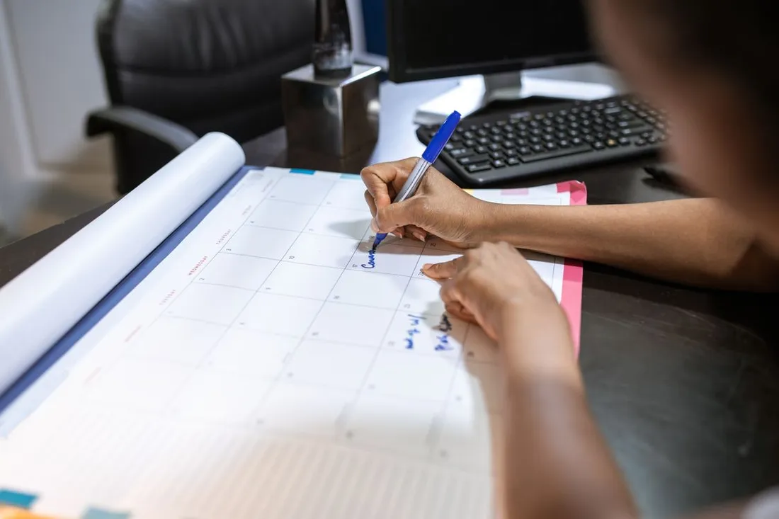 Main annotant au feutre bleu une case d'un grand calendrier mensuel en papier posé sur un bureau, devant un clavier et un écran