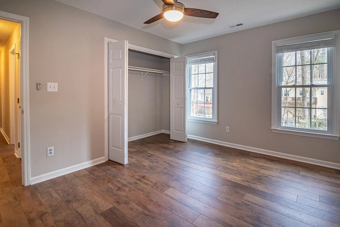 Chambre vide d'un logement non meublé : murs nus, parquet, ventilateur de plafond allumé, placard mural ouvert avec sa tringle et deux fenêtres donnant sur des arbres