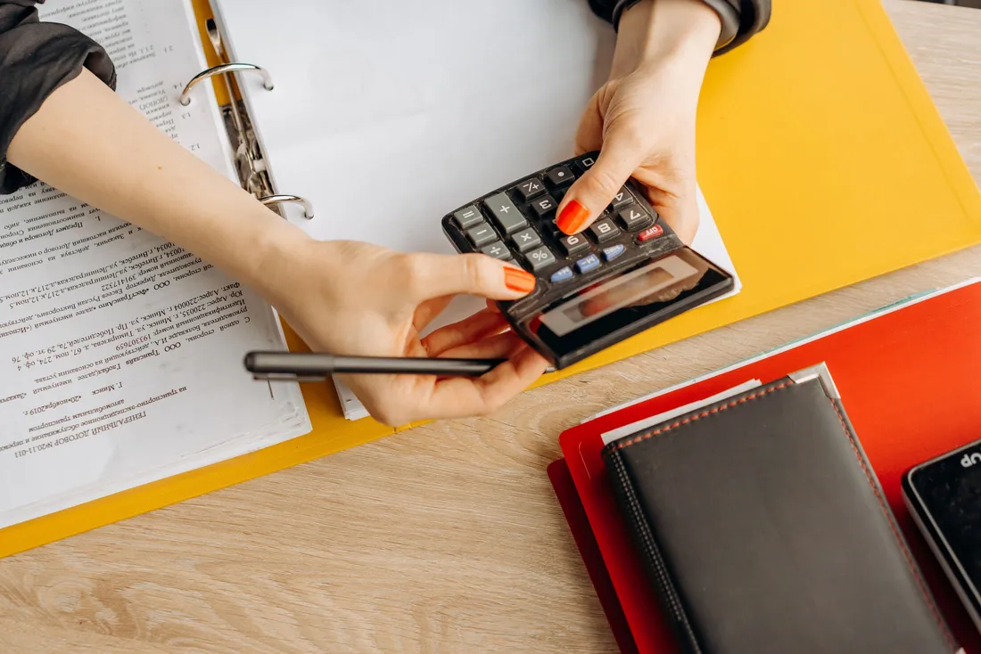 Mains d'une personne posant une calculatrice de poche sur un classeur ouvert contenant un contrat imprimé, à côté d'un stylo, de chemises cartonnées jaune et rouge et d'un carnet