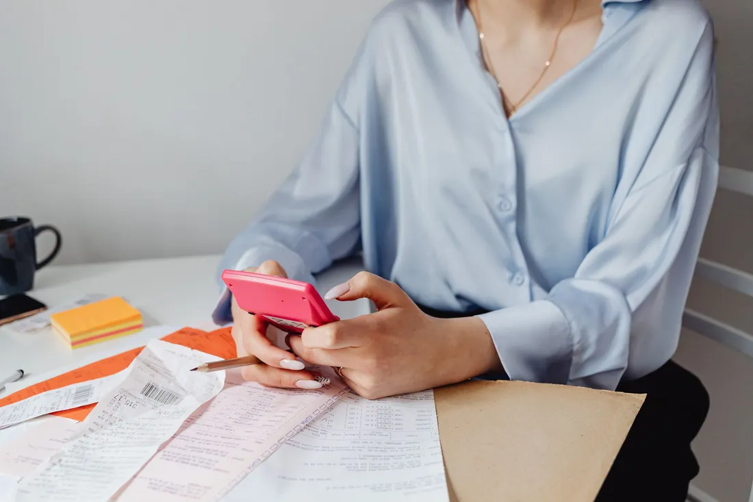 Une femme assise à un bureau tape sur une calculatrice, entourée de tickets, de relevés imprimés et d'une chemise cartonnée