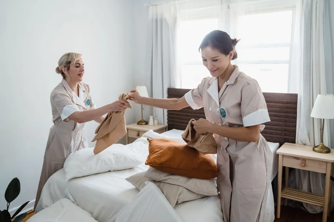 Deux employées d'entretien en tenue refont un lit et changent les taies d'oreiller dans une chambre claire