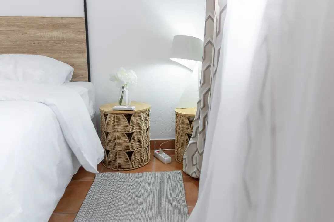 Chambre blanche de la Casa Belas Artes : lit à tête de bois clair, chevets en fibre tressée et lampe allumée derrière le voilage