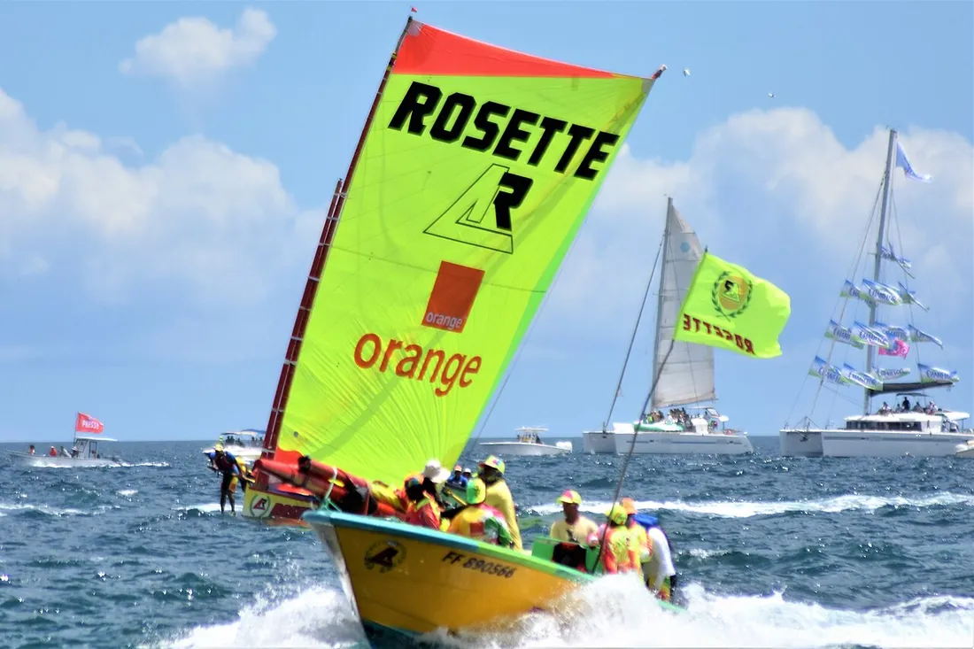 Yole ronde traditionnelle aux voiles colorées sponsorisées, propulsée par son équipage lors d'une course du Tour des Yoles en Martinique