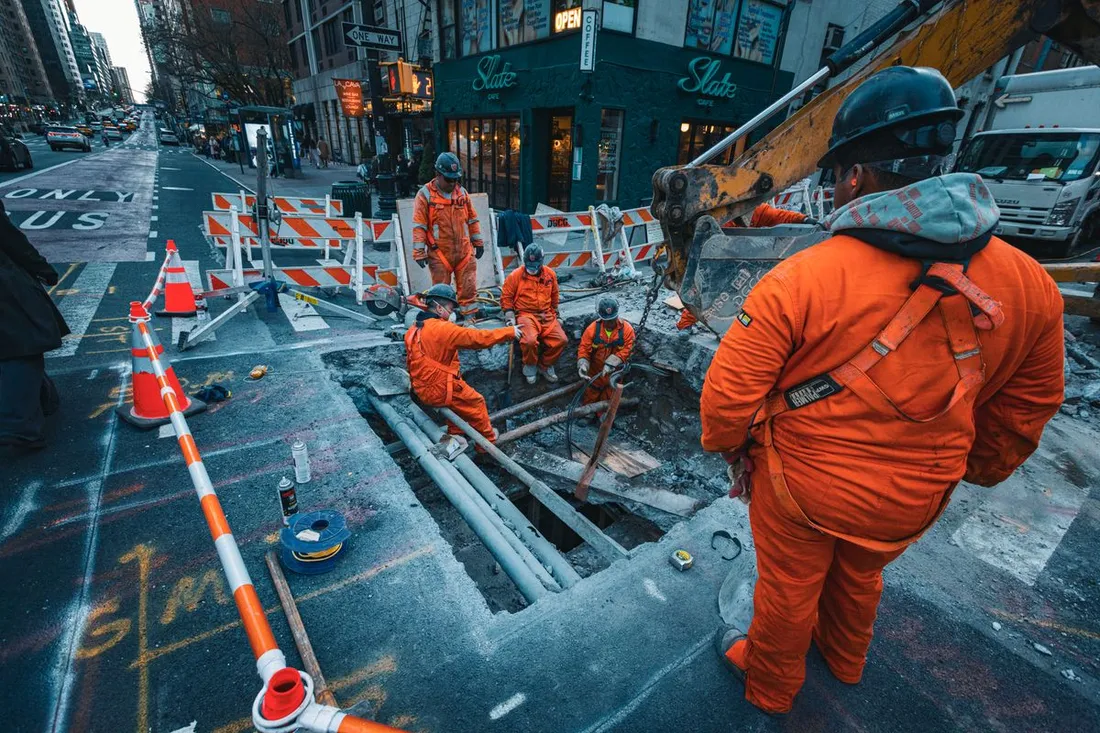 Chantier de voirie en pleine rue : une tranchée ouverte dans la chaussée, des ouvriers en combinaison orange autour, des barrières et des cônes qui neutralisent une voie