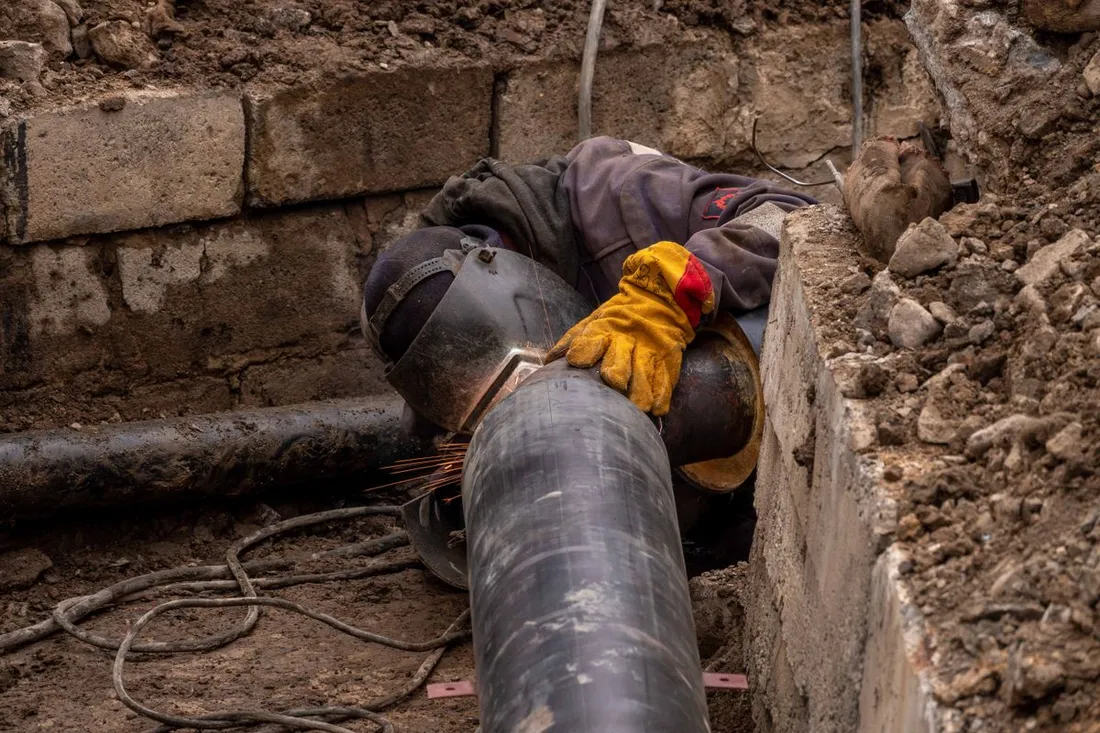 Au fond d'une tranchée, un ouvrier casqué de son masque de soudeur assemble deux tronçons de canalisation, des gerbes d'étincelles au point de soudure