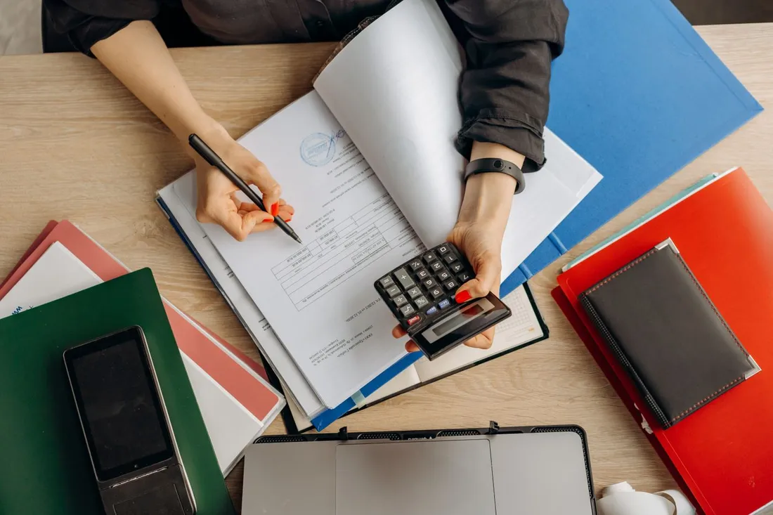 Gestionnaire calculant la TVA sur des factures et documents comptables avec une calculatrice, dossiers de location classés sur le bureau