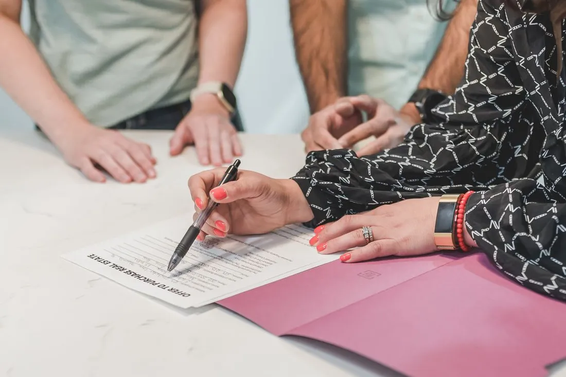 Mains signant au stylo une offre d'achat immobilier posée sur une table blanche, deux autres personnes debout autour du document