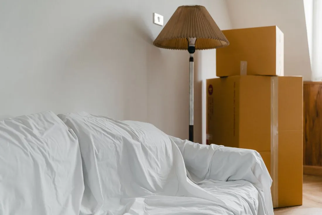 Canapé recouvert d'un drap blanc et cartons de déménagement empilés contre le mur d'une pièce vidée, à côté d'un lampadaire