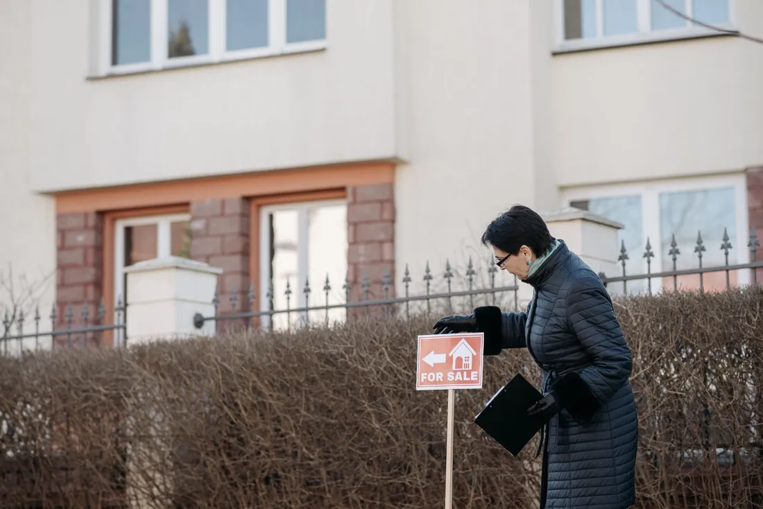 Une femme en manteau, un porte-documents sous le bras, installe un panneau « For Sale » devant une maison bordée d'une haie