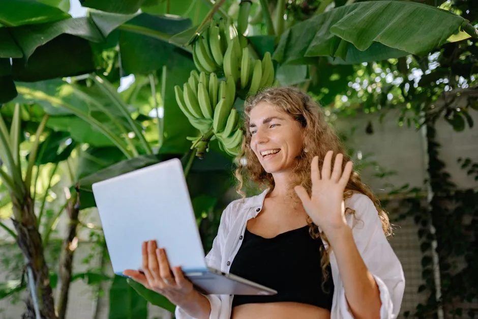 Femme souriante faisant un signe de la main devant l'écran de son ordinateur portable lors d'un appel vidéo, en extérieur sous des bananiers