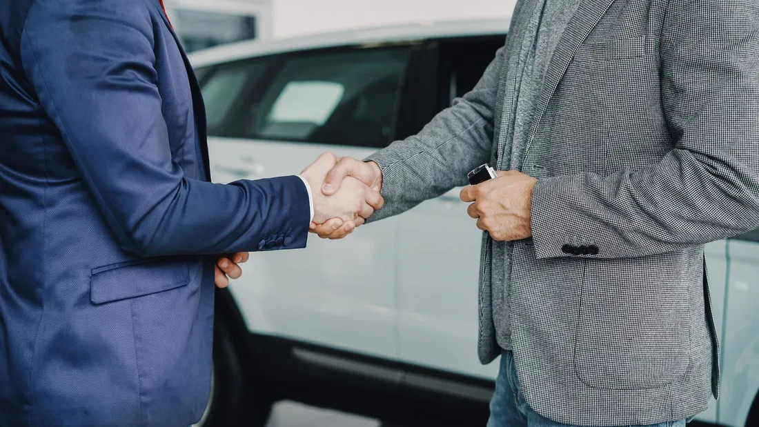 Poignée de main entre un vendeur en costume et un client devant une voiture blanche dans un hall de concession automobile
