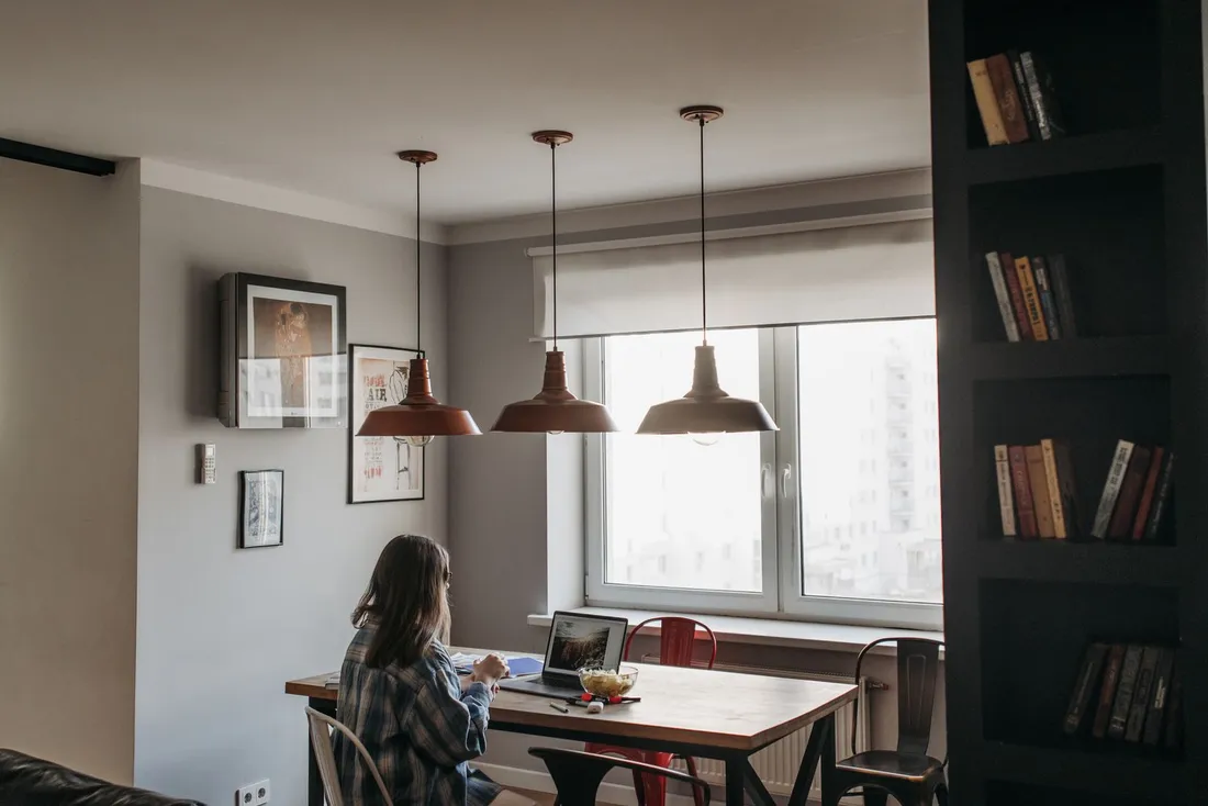 Voyageuse en télétravail installée à une table devant la fenêtre d'un appartement lumineux, ordinateur portable ouvert