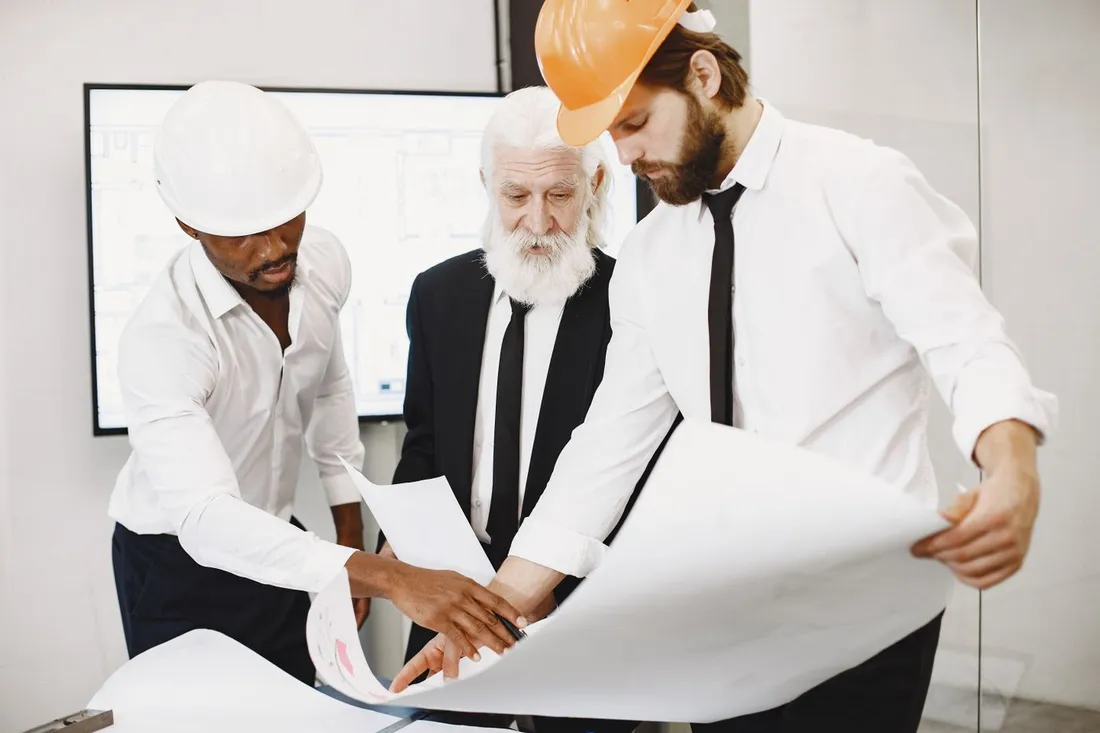 Trois hommes debout autour d'une table, deux d'entre eux coiffés d'un casque de chantier, penchés sur un grand plan de bâtiment déroulé